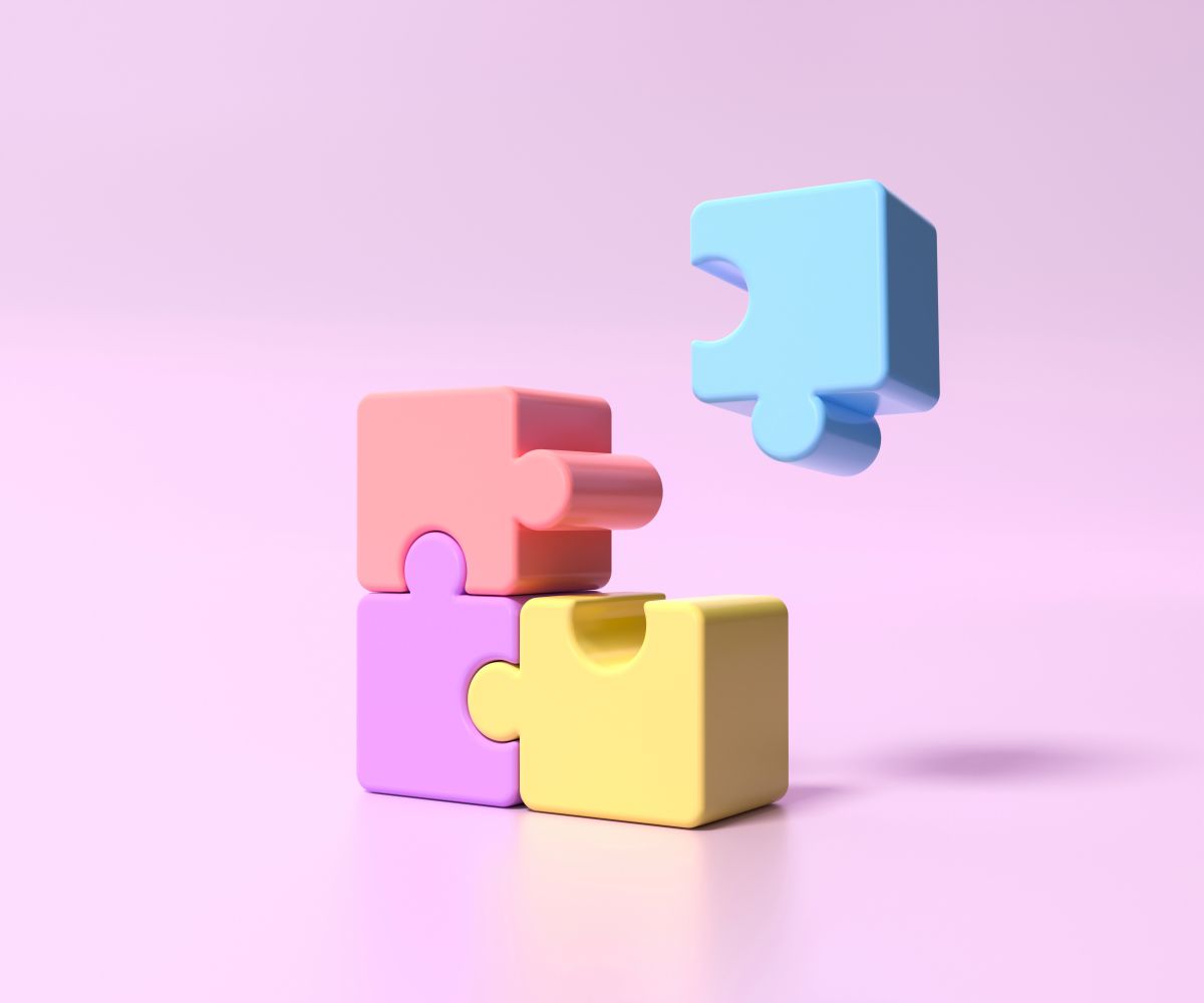 Colorful 3D puzzle pieces stacked and arranged against a pink background, with a blue piece floating above and pink, purple, and yellow pieces interlocking below, symbolizing problem-solving and teamwork.